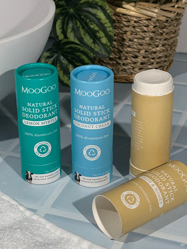 Three MooGoo solid deodorant tubes on blue bathroom tiles: turquoise Lemon Myrtle, blue Coconut Cream and tan Oats and Honey, with the tan stick open and its cap lying in front.
