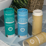 Three MooGoo solid deodorant tubes on blue bathroom tiles: turquoise Lemon Myrtle, blue Coconut Cream and tan Oats and Honey, with the tan stick open and its cap lying in front.