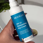 Hand with lilac nail polish holding an uncapped blue-labelled MooGoo SPF 15 Moisturising Day Cream pump bottle beside a bathroom basin.