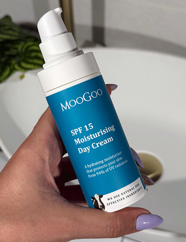 Hand with lilac nail polish holding an uncapped blue-labelled MooGoo SPF 15 Moisturising Day Cream pump bottle beside a bathroom basin.