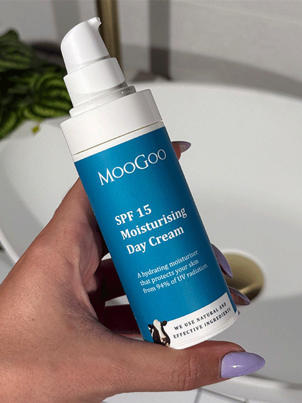 Hand with lilac nail polish holding an uncapped blue-labelled MooGoo SPF 15 Moisturising Day Cream pump bottle beside a bathroom basin.