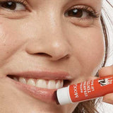 Smiling woman holding MooGoo Strawberry Tinted Lip Balm beside her lips, showing the warm red tint.