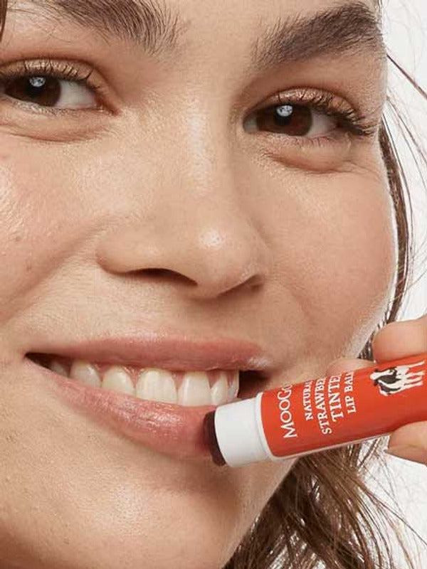 Smiling woman holding MooGoo Strawberry Tinted Lip Balm beside her lips, showing the warm red tint.