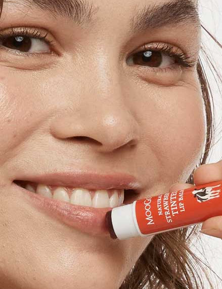 A woman with clear skin and natural makeup smiles as she holds MooGoo Lip Balm - Strawberry Tinted 5g near her lips, preparing to apply the red-tinted balm. Her face is close to the camera.