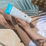 Person seated outdoors holding a white-and-blue MooGoo Clear Zinc sunscreen tube over their bare legs, with sunscreen on one knee and a straw bag beside them.
