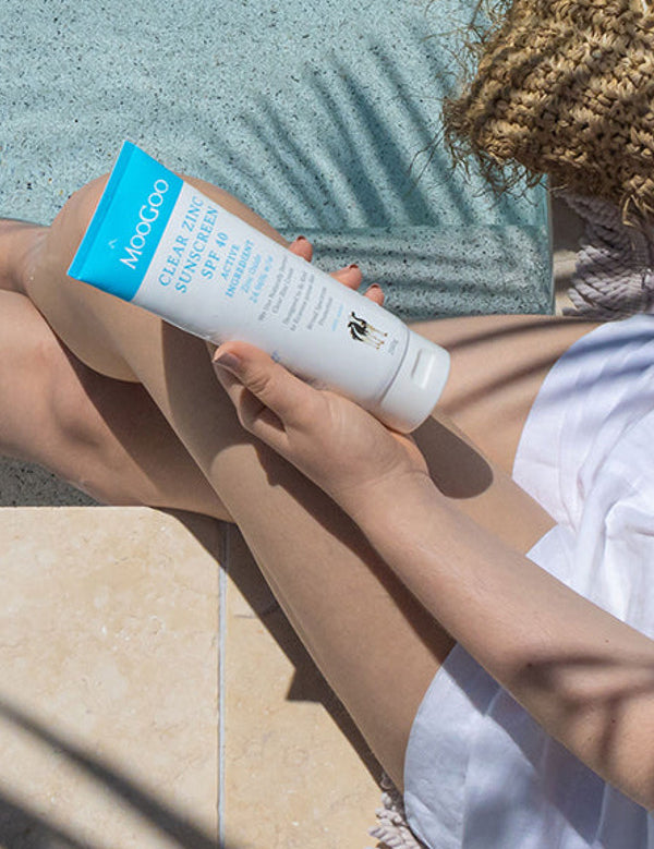 Person seated outdoors holding a white-and-blue MooGoo Clear Zinc sunscreen tube over their bare legs, with sunscreen on one knee and a straw bag beside them.
