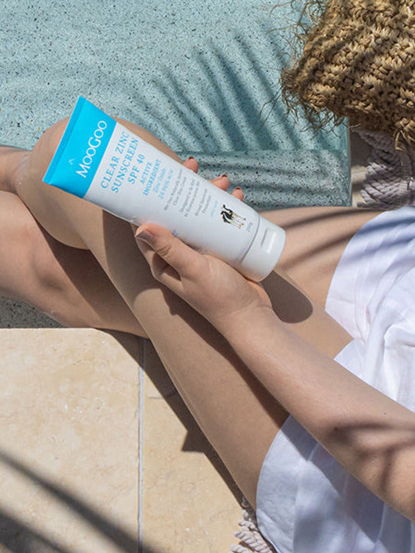 Person seated outdoors holding a white-and-blue MooGoo Clear Zinc sunscreen tube over their bare legs, with sunscreen on one knee and a straw bag beside them.