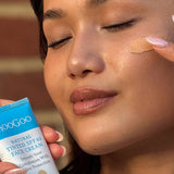 Close-up of a person with closed eyes dabbing beige tinted cream onto their cheek while holding a MooGoo Tinted SPF 40 Face Cream tube.