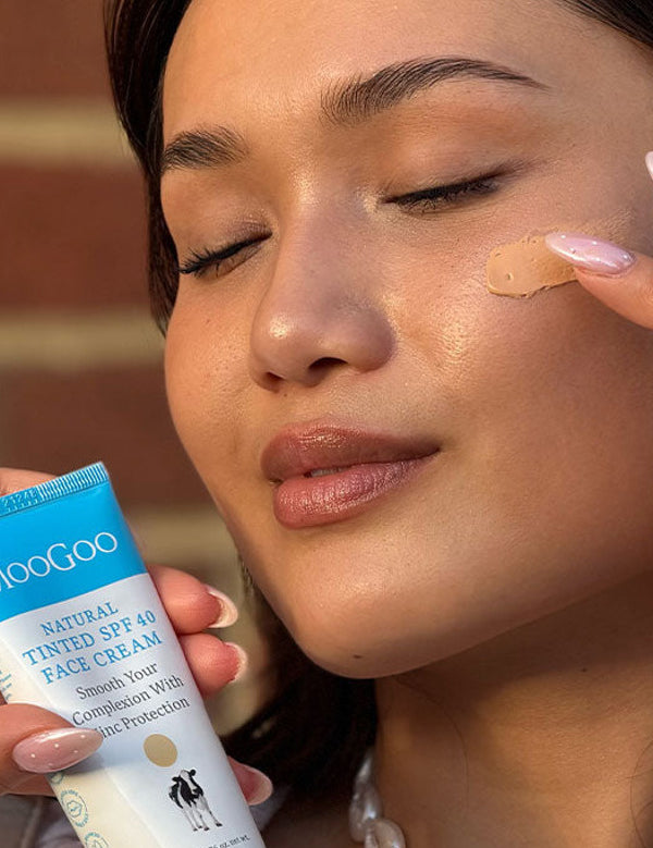 Close-up of a person with closed eyes dabbing beige tinted cream onto their cheek while holding a MooGoo Tinted SPF 40 Face Cream tube.