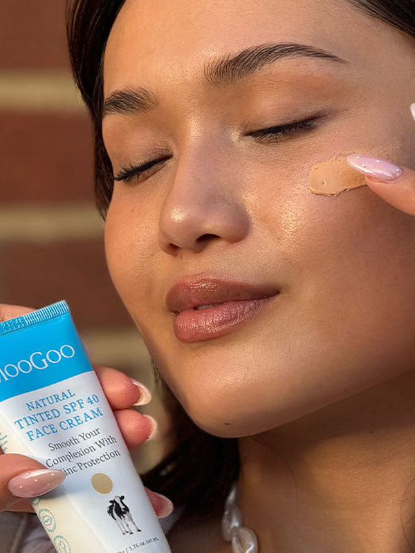 Close-up of a person with closed eyes dabbing beige tinted cream onto their cheek while holding a MooGoo Tinted SPF 40 Face Cream tube.