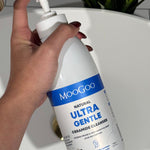 Hand holding a large MooGoo Ultra Gentle Cleanser with Ceramides pump bottle beside a bathroom basin.