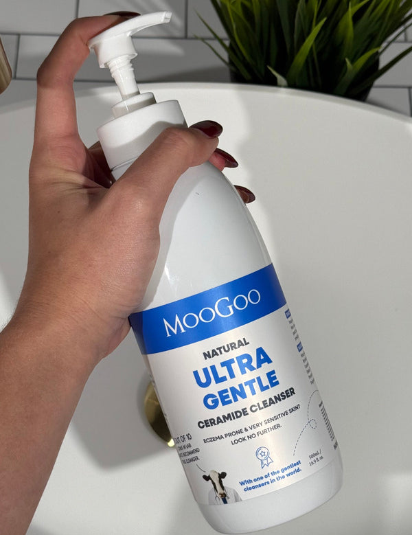 Hand holding a large MooGoo Ultra Gentle Cleanser with Ceramides pump bottle beside a bathroom basin.