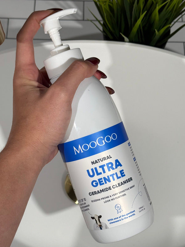 Hand holding a large MooGoo Ultra Gentle Cleanser with Ceramides pump bottle beside a bathroom basin.