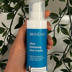 Hand holding a blue-labelled MooGoo Ultra Hydrating Face Cream pump bottle with its clear cap against trailing green leaves.