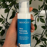 Hand holding a blue-labelled MooGoo Ultra Hydrating Face Cream pump bottle with its clear cap against trailing green leaves.