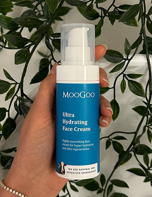 Hand holding a blue-labelled MooGoo Ultra Hydrating Face Cream pump bottle with its clear cap against trailing green leaves.