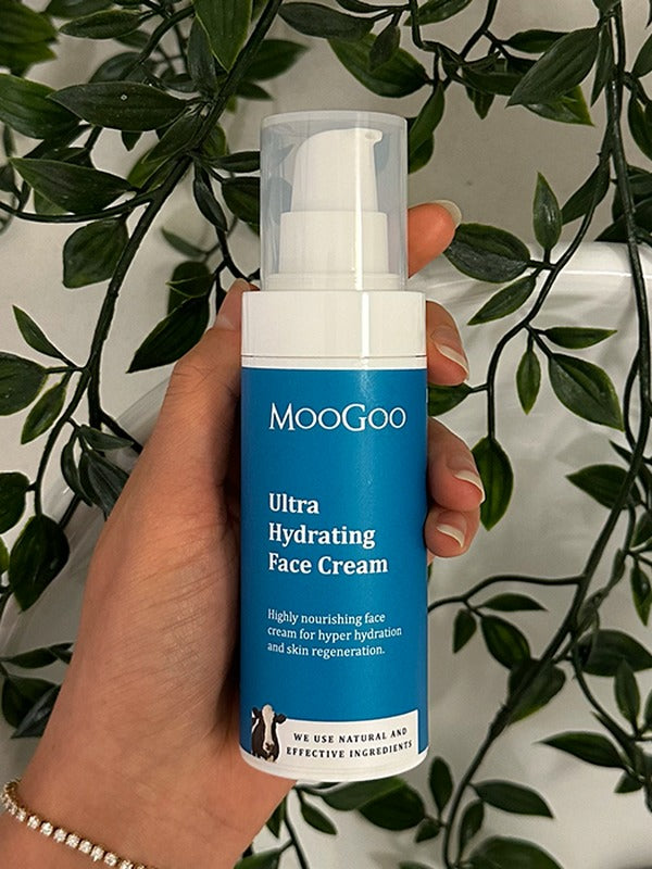 Hand holding a blue-labelled MooGoo Ultra Hydrating Face Cream pump bottle with its clear cap against trailing green leaves.