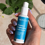 Hand with lilac nails holding an uncapped blue-labelled MooGoo Vitamin C Brightening Active Serum pump bottle beside a leafy plant and mirror.
