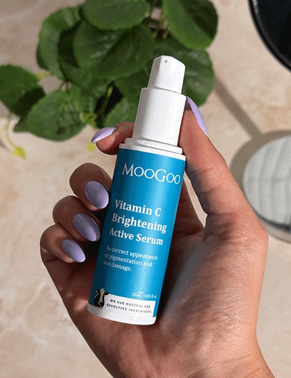 Hand with lilac nails holding an uncapped blue-labelled MooGoo Vitamin C Brightening Active Serum pump bottle beside a leafy plant and mirror.