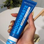 Hand with yellow nails holding a blue MooGoo Whitening Peppermint Moothpaste tube beside a wooden toothbrush over a white basin.