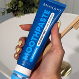 Hand with yellow nails holding a blue MooGoo Whitening Peppermint Moothpaste tube beside a wooden toothbrush over a white basin.