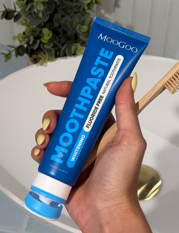 Hand with yellow nails holding a blue MooGoo Whitening Peppermint Moothpaste tube beside a wooden toothbrush over a white basin.