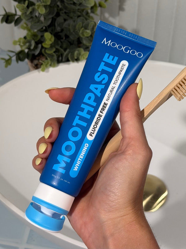 Hand with yellow nails holding a blue MooGoo Whitening Peppermint Moothpaste tube beside a wooden toothbrush over a white basin.