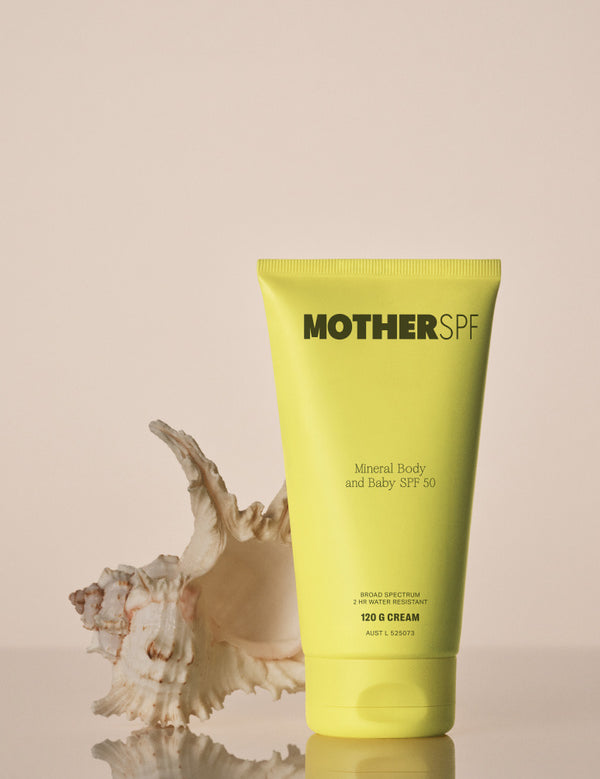 MOTHER SPF Mineral Body and Baby SPF 50 120g yellow tube beside a seashell on a reflective surface with a warm beige background.