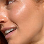 Mukti Golden Glow Bronzing Drops worn on the face: close-up of a model's cheek showing the golden finish in the brand's two-layer application photo.