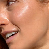 Mukti Golden Glow Bronzing Drops worn on the face: close-up of a model's cheek showing the golden finish in the brand's two-layer application photo.