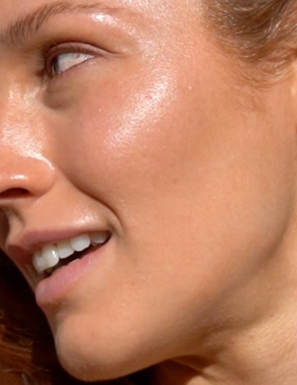 Mukti Golden Glow Bronzing Drops worn on the face: close-up of a model's cheek showing the golden finish in the brand's two-layer application photo.