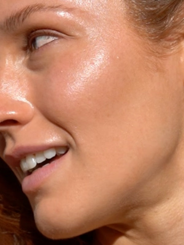Mukti Golden Glow Bronzing Drops worn on the face: close-up of a model's cheek showing the golden finish in the brand's two-layer application photo.