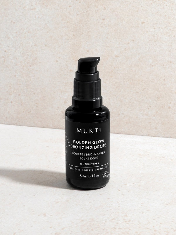 Mukti Golden Glow Bronzing Drops 30ml black pump bottle standing on a pale stone surface with its front label visible.