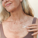 Mature woman smiling while applying Mukti Age Defiance Body Serum to her shoulder.