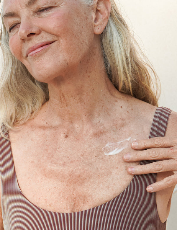 Mature woman smiling while applying Mukti Age Defiance Body Serum to her shoulder.
