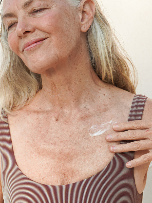 Mature woman smiling while applying Mukti Age Defiance Body Serum to her shoulder.