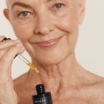 Silver-haired woman applying Mukti Antioxidant Facial Oil with a glass dropper while holding the bottle.