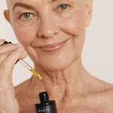 Silver-haired woman applying Mukti Antioxidant Facial Oil with a glass dropper while holding the bottle.