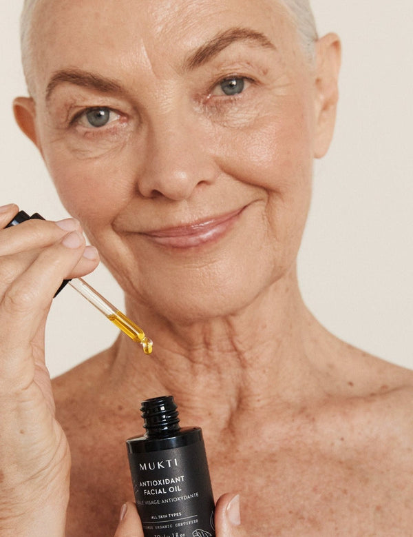 Silver-haired woman applying Mukti Antioxidant Facial Oil with a glass dropper while holding the bottle.