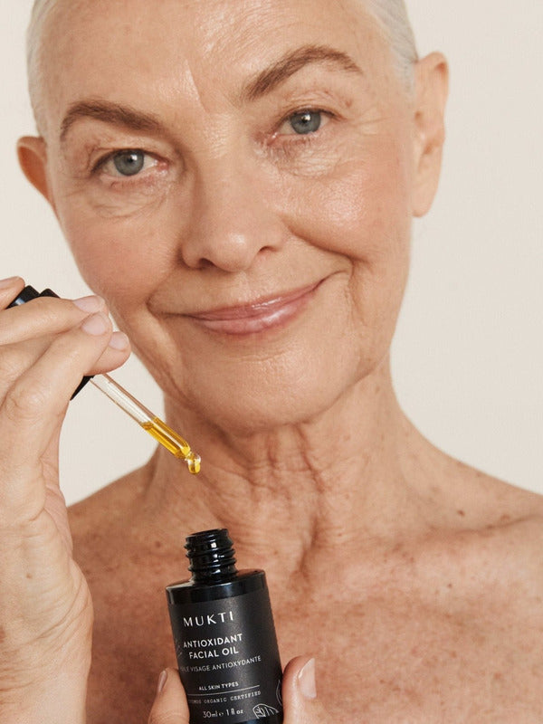 Silver-haired woman applying Mukti Antioxidant Facial Oil with a glass dropper while holding the bottle.