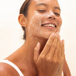 Woman smiling while massaging Mukti Balancing Cleansing Gel over her cheeks against a bright neutral background.