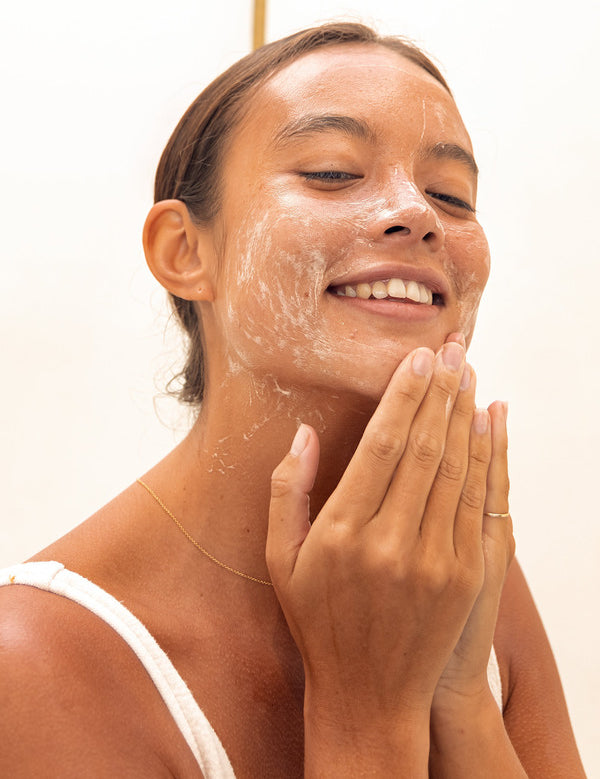 Woman smiling while massaging Mukti Balancing Cleansing Gel over her cheeks against a bright neutral background.