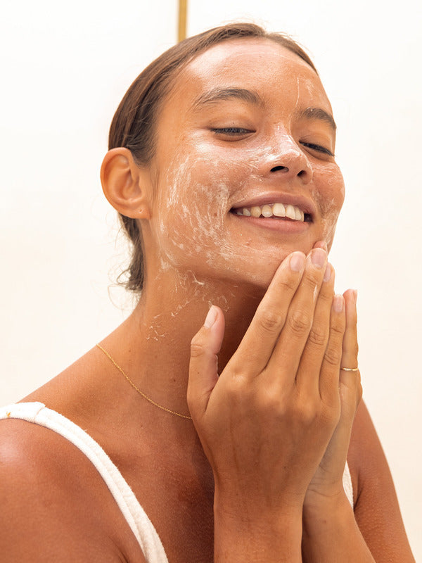 Woman smiling while massaging Mukti Balancing Cleansing Gel over her cheeks against a bright neutral background.