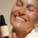 Smiling woman holding Mukti Bioactive Renewal Day Serum beside her face in warm natural light.