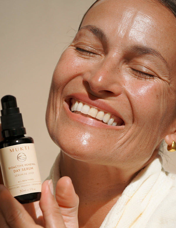 Smiling woman holding Mukti Bioactive Renewal Day Serum beside her face in warm natural light.