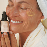 Smiling woman in a white robe holding Mukti Bioactive Renewal Night Serum beside her face, with a serum swatch on her cheek.