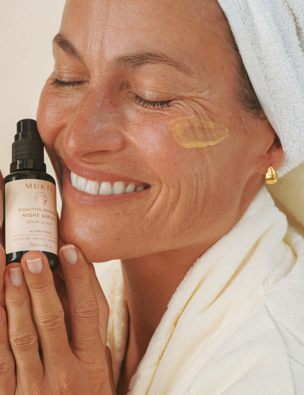 Smiling woman in a white robe holding Mukti Bioactive Renewal Night Serum beside her face, with a serum swatch on her cheek.