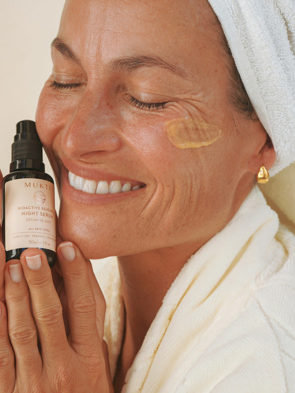 Smiling woman in a white robe holding Mukti Bioactive Renewal Night Serum beside her face, with a serum swatch on her cheek.