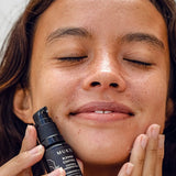 Woman with closed eyes holding Mukti Blemish Control Acne Spot Treatment 15ml beside her face while applying skincare.