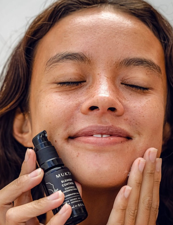 Woman with closed eyes holding Mukti Blemish Control Acne Spot Treatment 15ml beside her face while applying skincare.